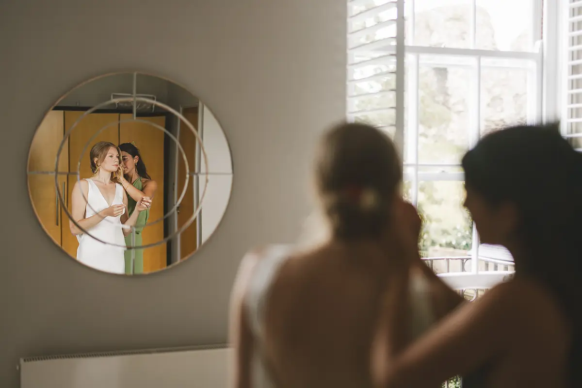 Bride and her bridesmaid in green reflected together in a round mirror during wedding preparations