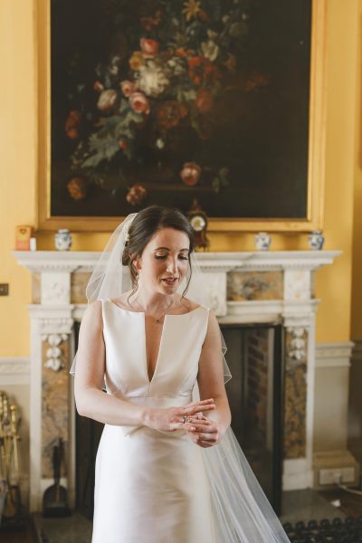 Elegant bride in white gown with veil standing before an ornate marble fireplace and floral oil painting inside Smedmore House