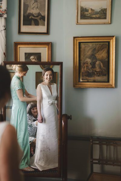 Smiling bride seen in a full-length mirror as her bridesmaid fastens her dress in a room filled with framed artwork at Smedmore House