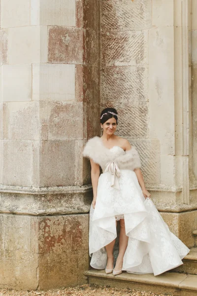 Bride in a high-low wedding gown and fur wrap leaning against the stone exterior of Highcliffe Castle