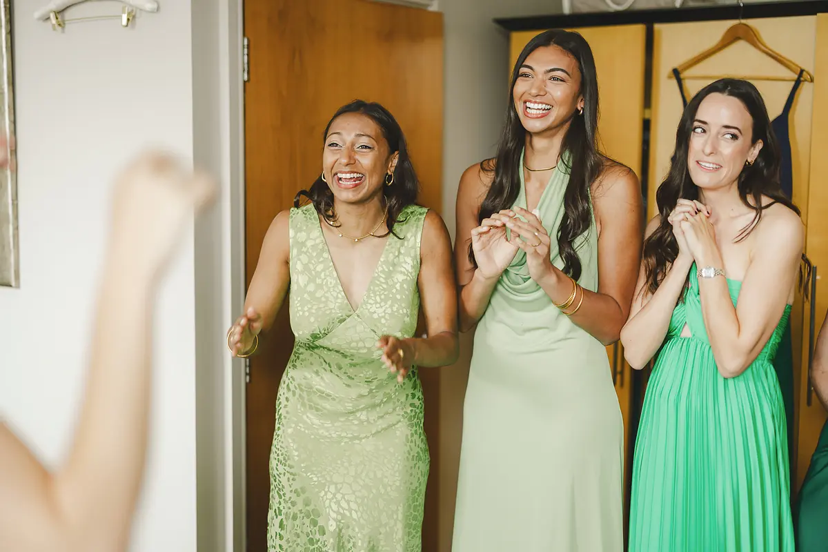 Three bridesmaids in green dresses laughing and clapping joyfully as the bride is fully ready