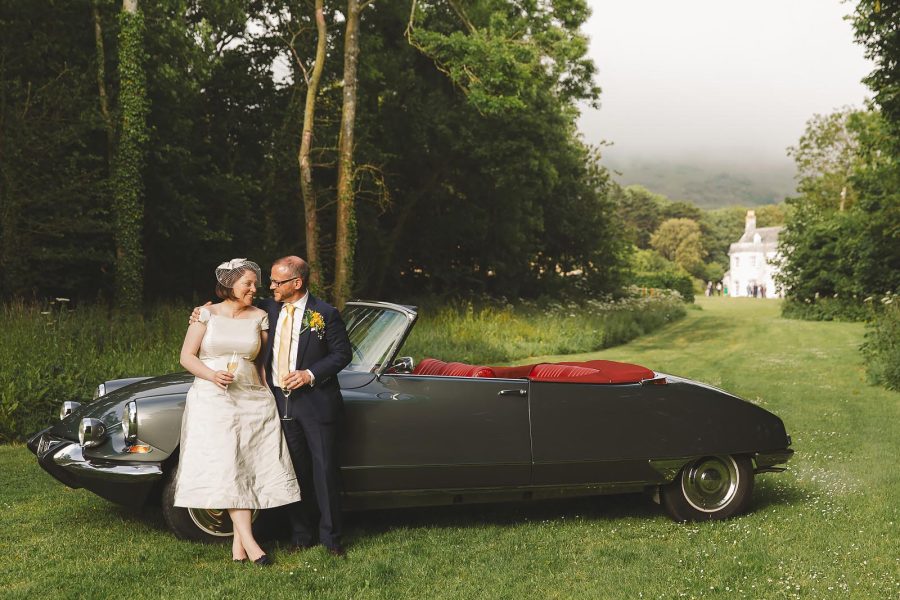 Bride and groom leaning against a vintage Citroen DS convertible in the grounds at Smedmore House, misty hills behind