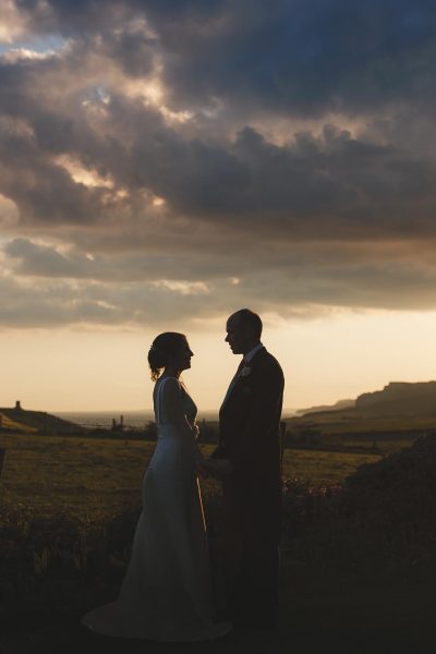 Silhouette of bride and groom facing each other against a dramatic Dorset sunset sky with Kimmeridge Bay visible in the distance