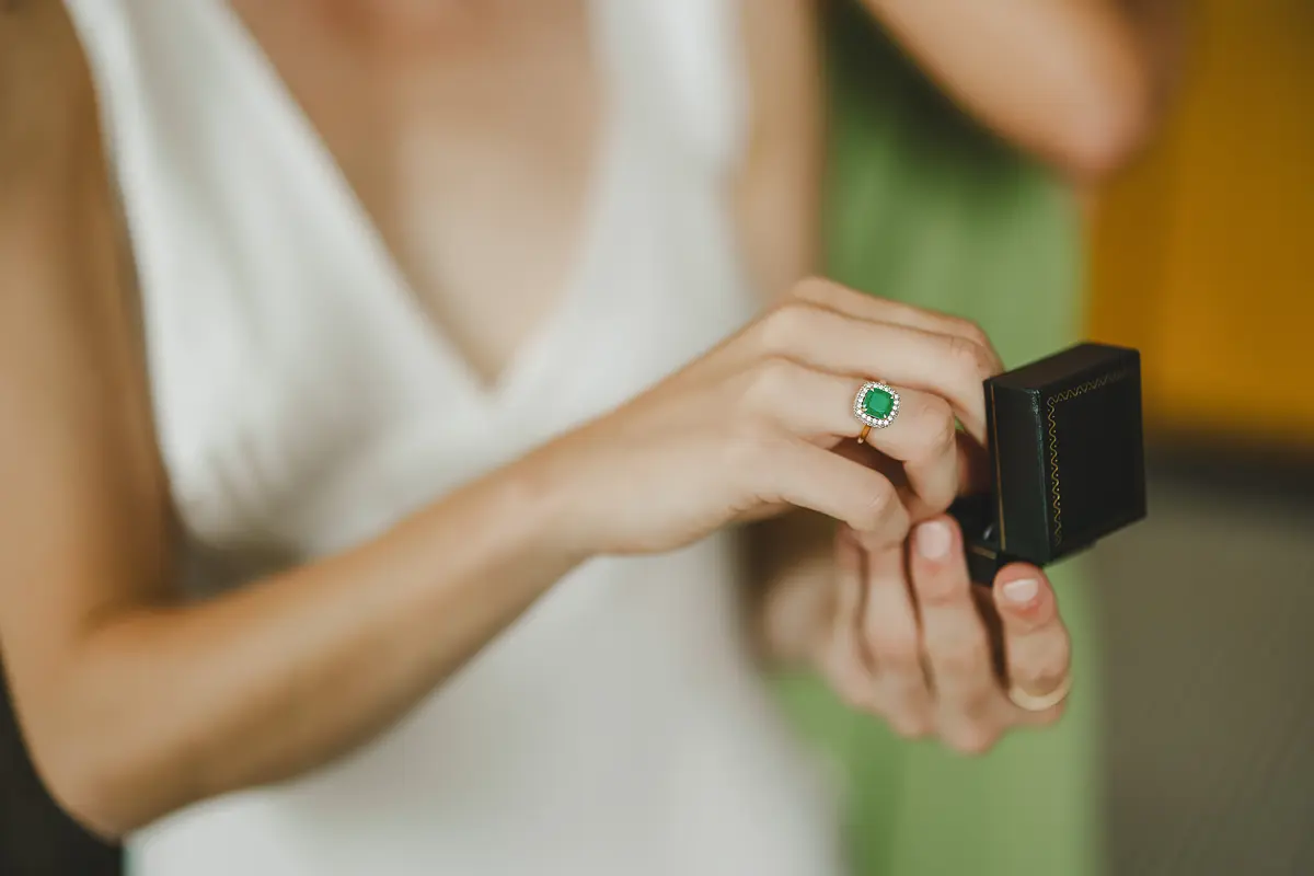 Bride's hand over a dark ring box, emerald and diamond engagement ring clearly visible