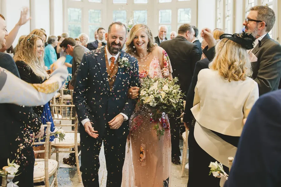 Bride in a colourful embroidered floral gown and groom walking through confetti in the Wintergarden at Highcliffe Castle