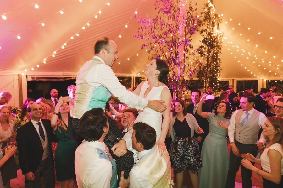 Groom being lifted on guests' shoulders on the dance floor while bride laughs under warm marquee lights at Smedmore House