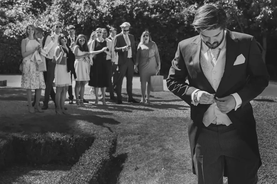 Groom adjusting his cufflinks in the Highcliffe Castle parterre garden with guests mingling in the background