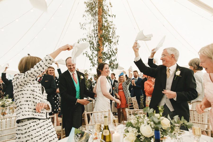 Bride and groom entering the marquee reception at Smedmore House to cheering guests waving white napkins under fairy lights