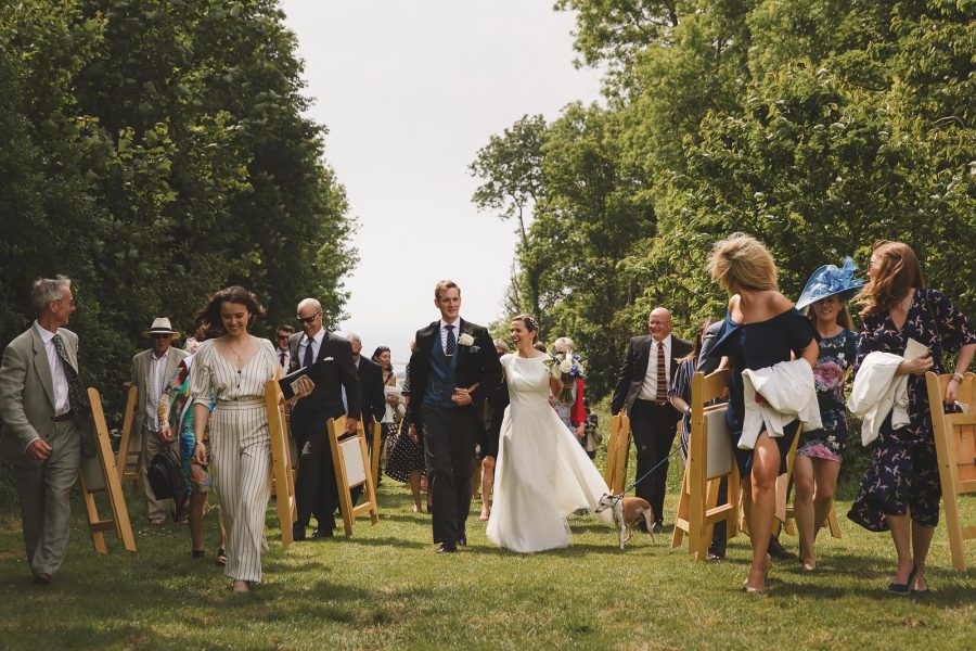 Newlywed couple walking back up the aisle after their outdoor ceremony at Smedmore House accompanied by their dog