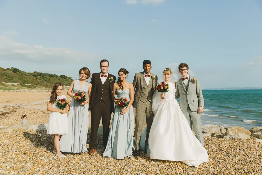 Wedding party group portrait on the pebble beach near Highcliffe Castle with the blue sea behind them