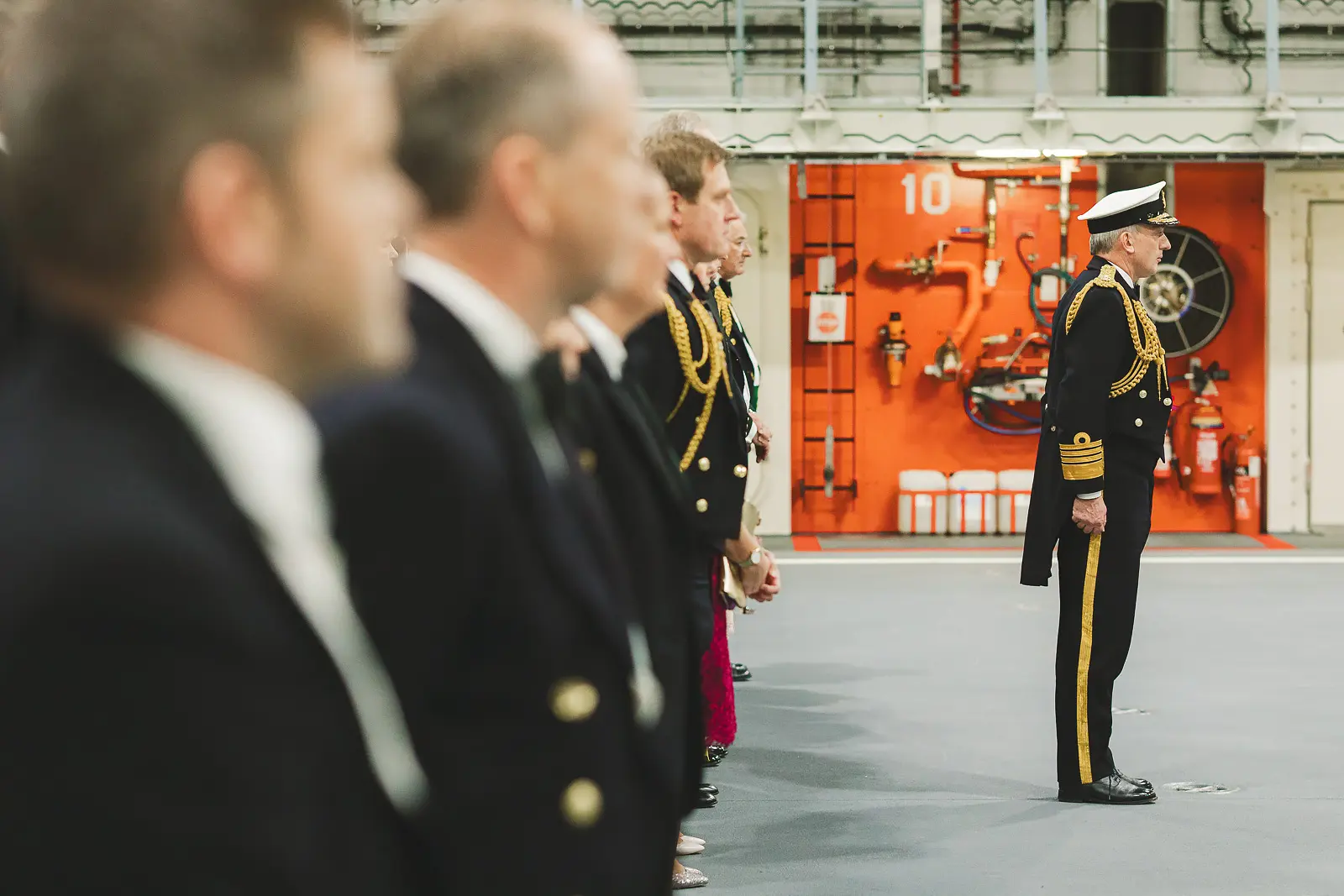 Senior admiral in full uniform facing line of officers and guests at attention during formal proceedings, fire safety cabinet visible aboard ship