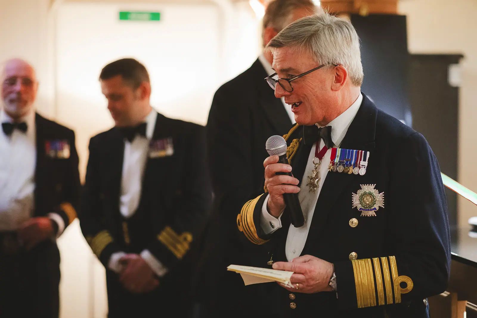 Senior admiral with extensive medals and decorations holding microphone delivering speech at formal military dinner, officers behind