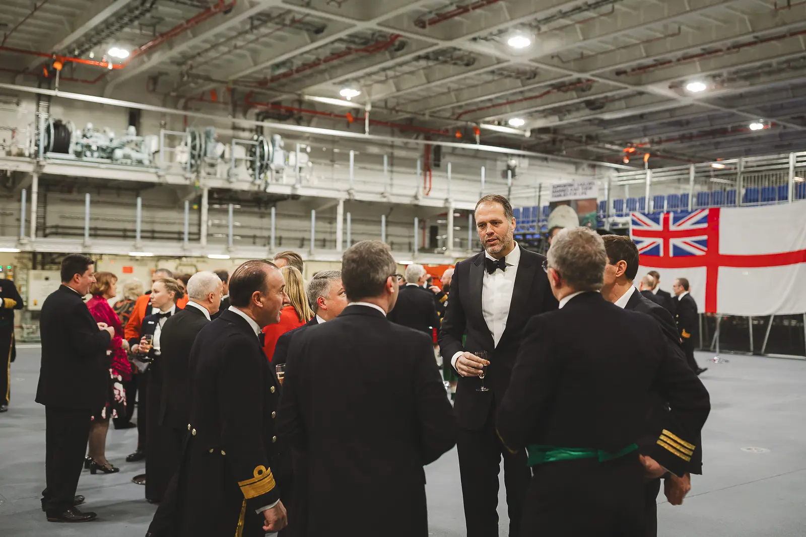 Wide view of hangar deck transformed for formal evening reception with naval officers and civilian guests in black tie, White Ensign displayed