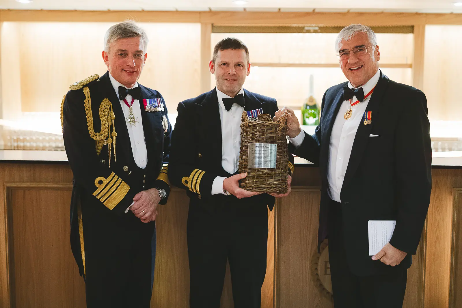 Senior naval officer, award recipient holding wicker hamper, and decorated civilian posing at formal dinner charity auction or awards presentation