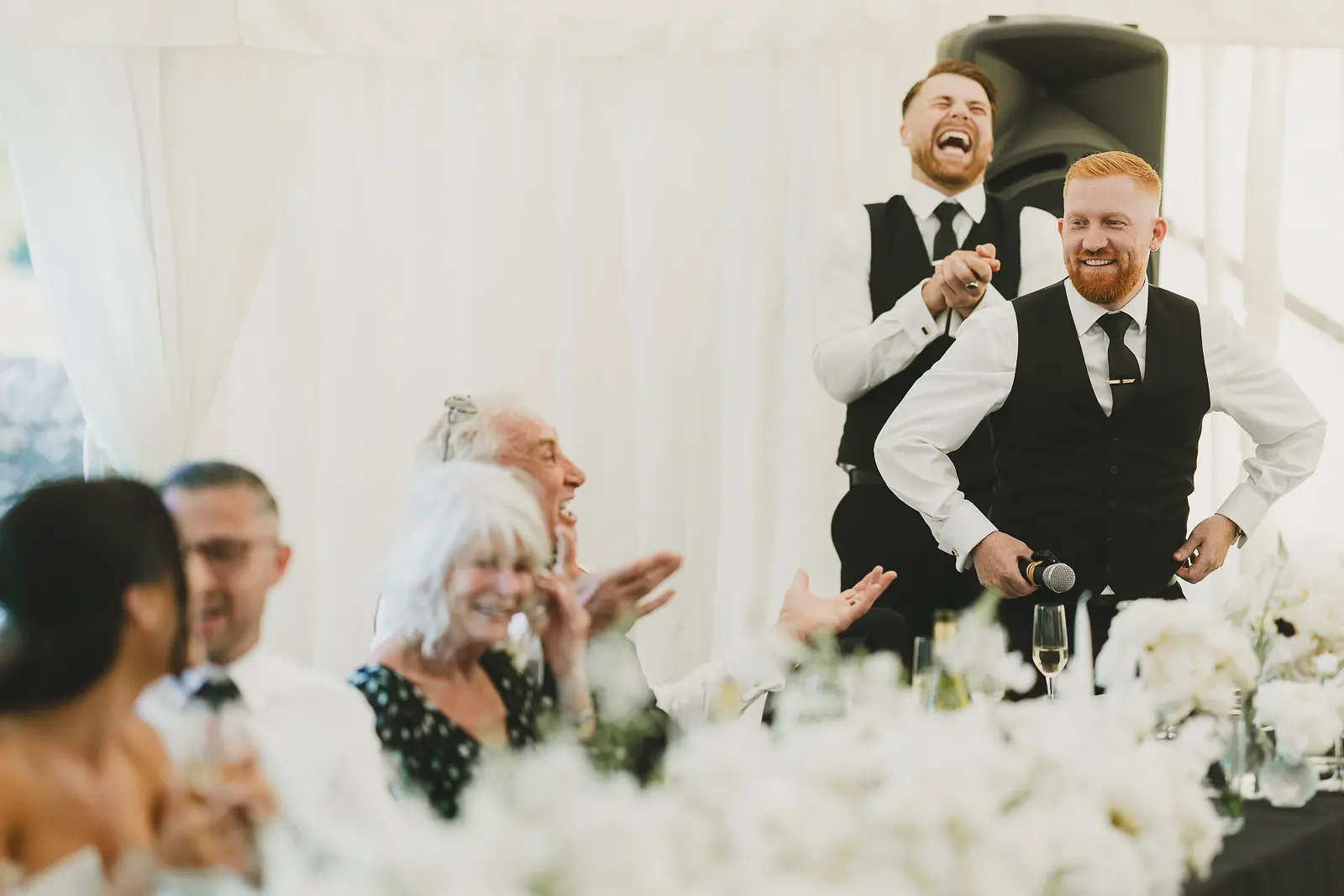 Best man delivering his speech to laughter from the top table in the marquee, guests enjoying the humour