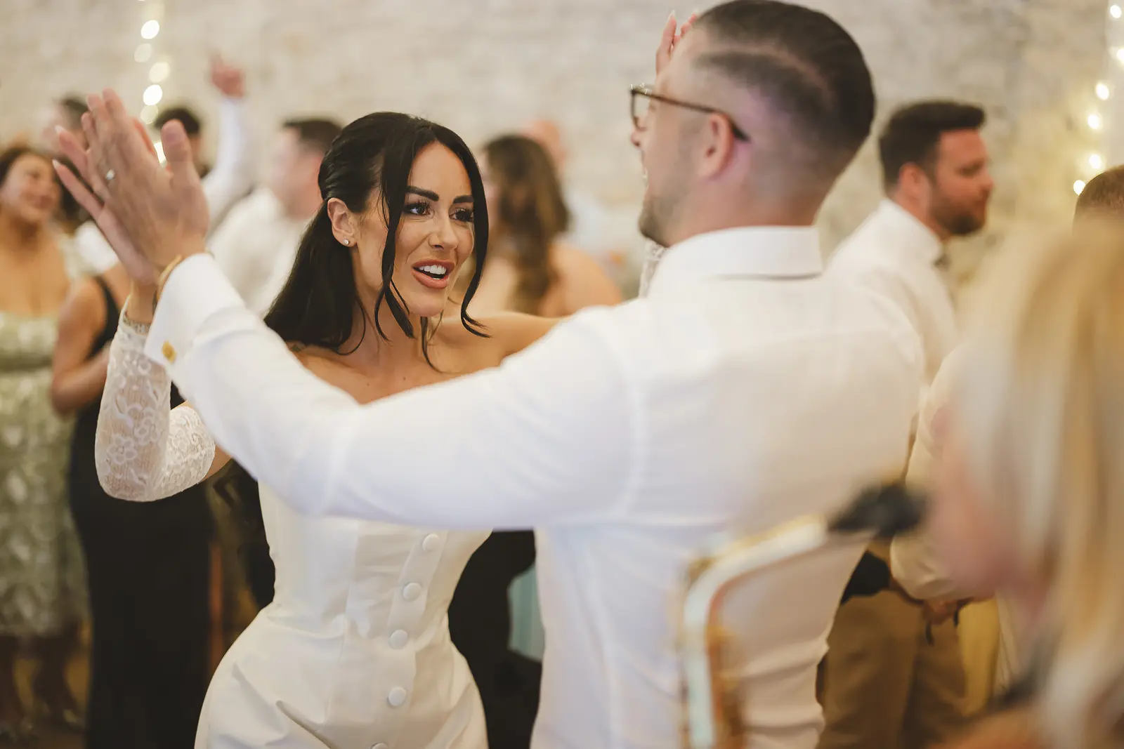 Bride in white mini dress with lace sleeves dancing energetically with the groom in the stone barn evening reception, guests dancing all around