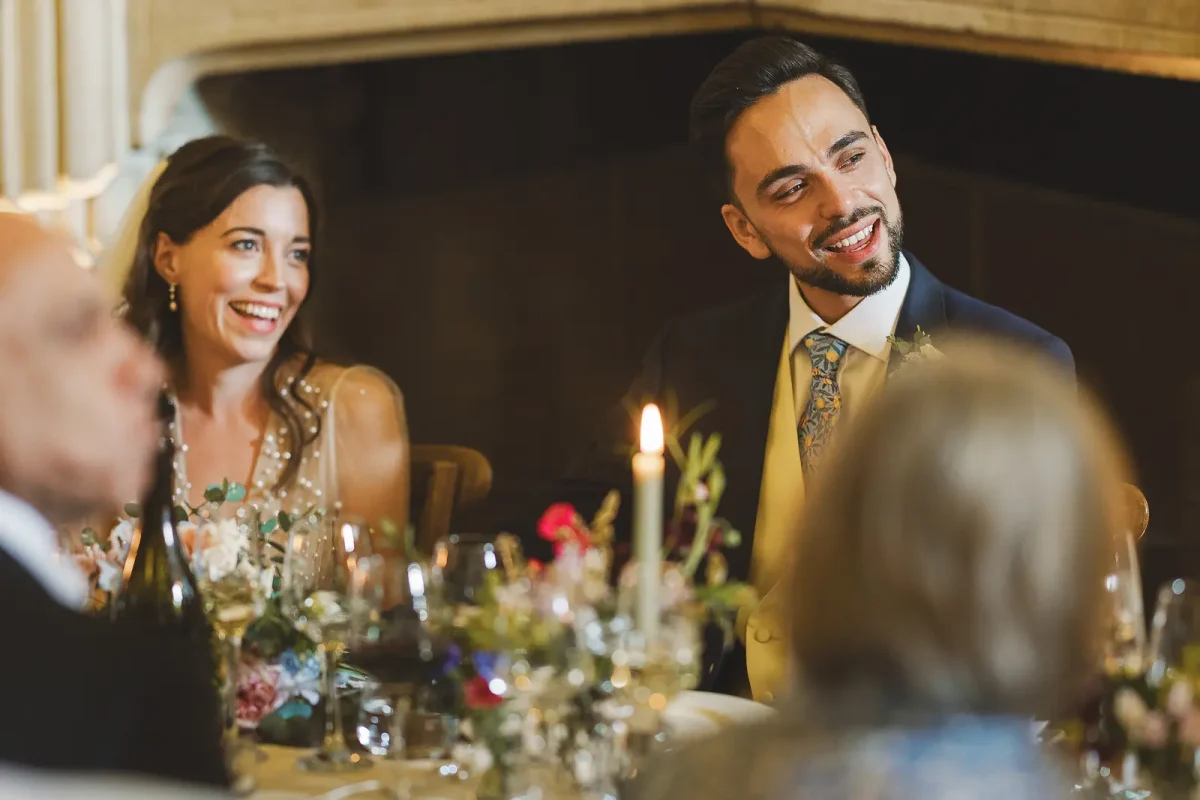 Bride and groom beaming with laughter at the candlelit top table with flowers and the stone fireplace behind during the wedding reception