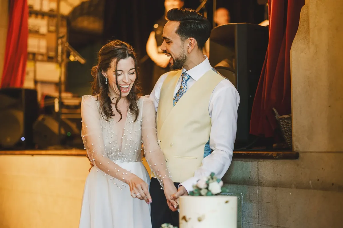 Bride and groom laughing together as they cut the wedding cake in the warm light of the tithe barn at Hinton St Mary Estate