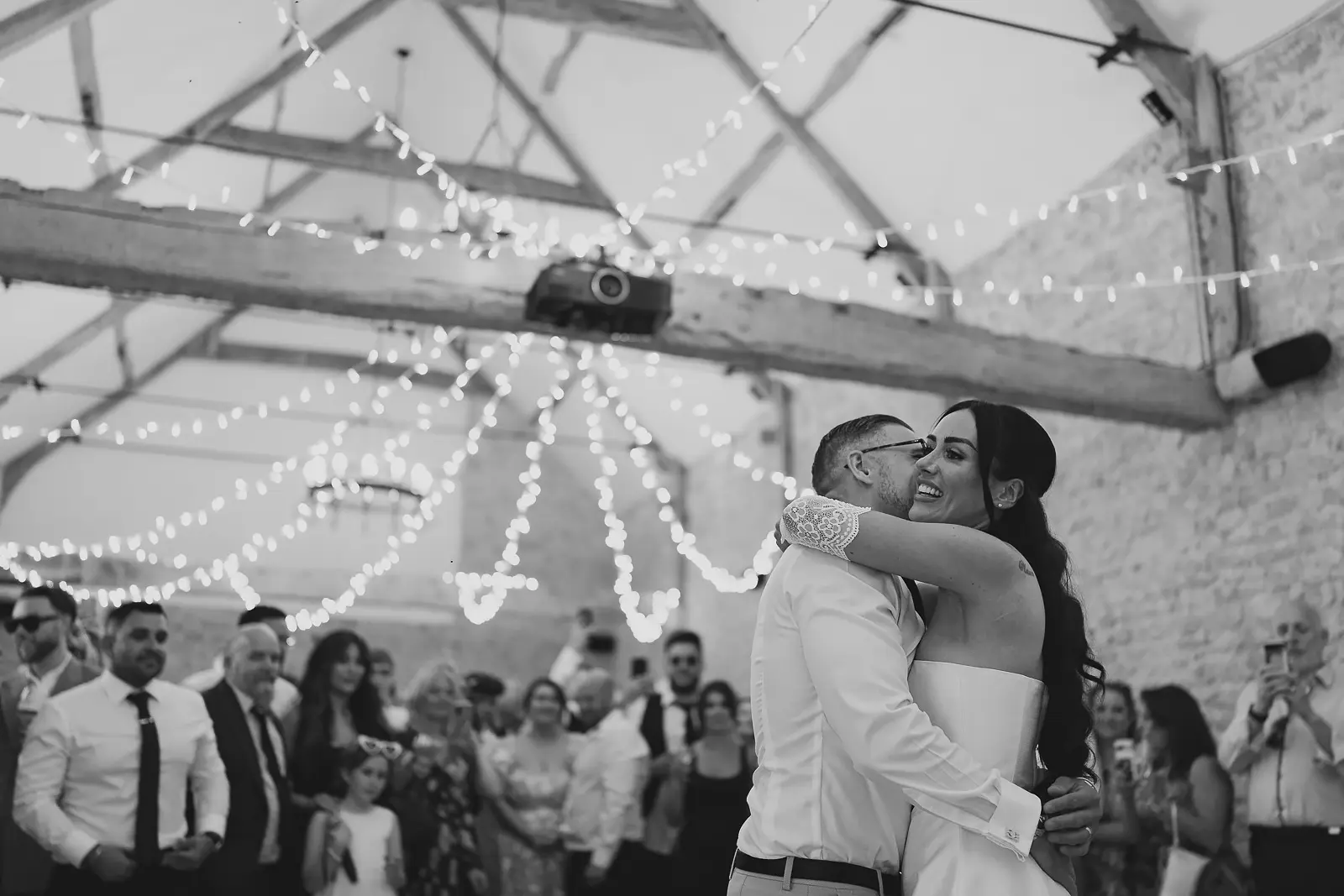 Black and white photograph of bride and groom sharing their first dance in the stone barn, fairy lights draping overhead, intimate and romantic