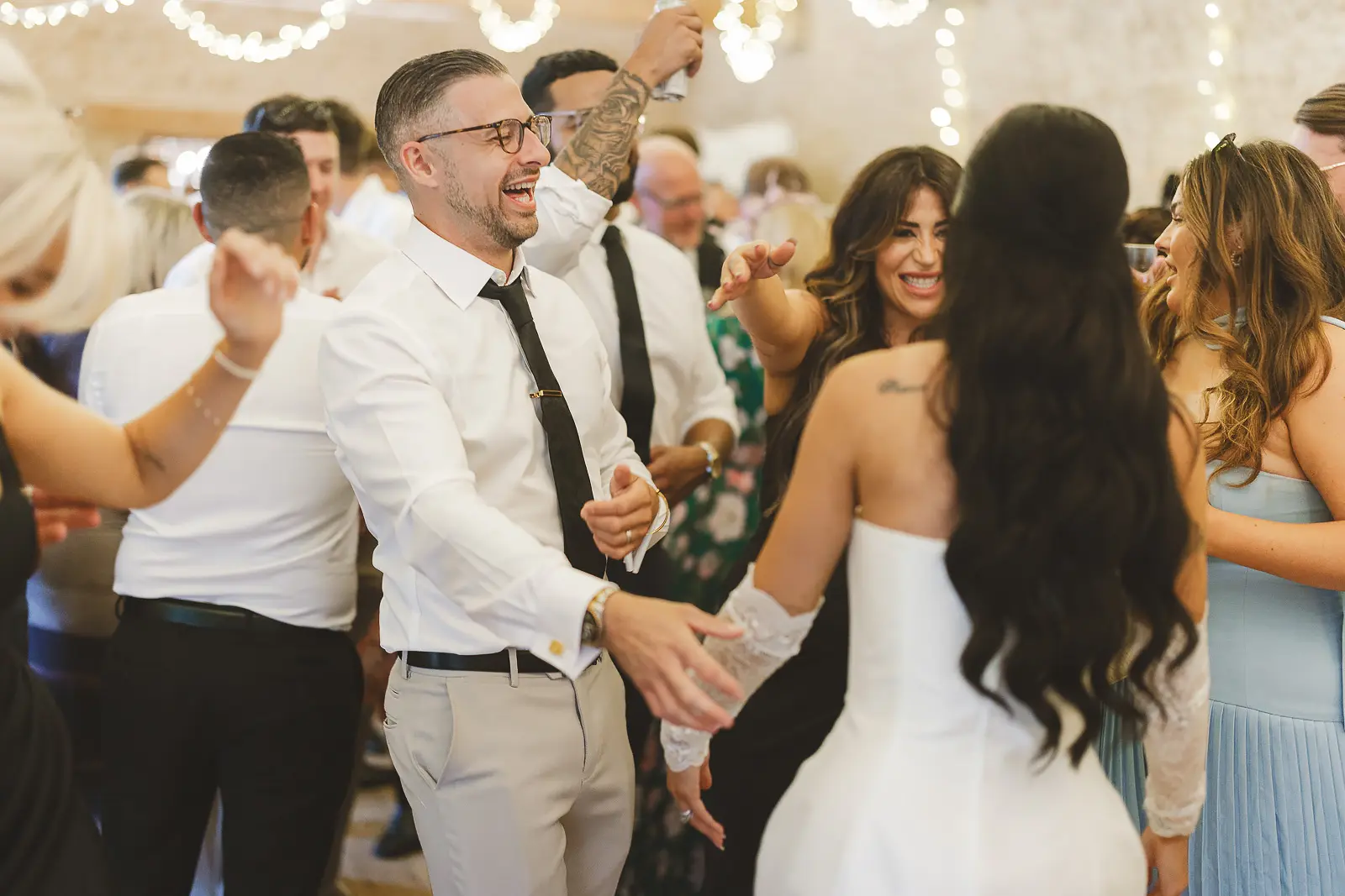 Bride and groom laughing together on the dance floor at the stone barn evening reception, fairy lights above and guests dancing around them