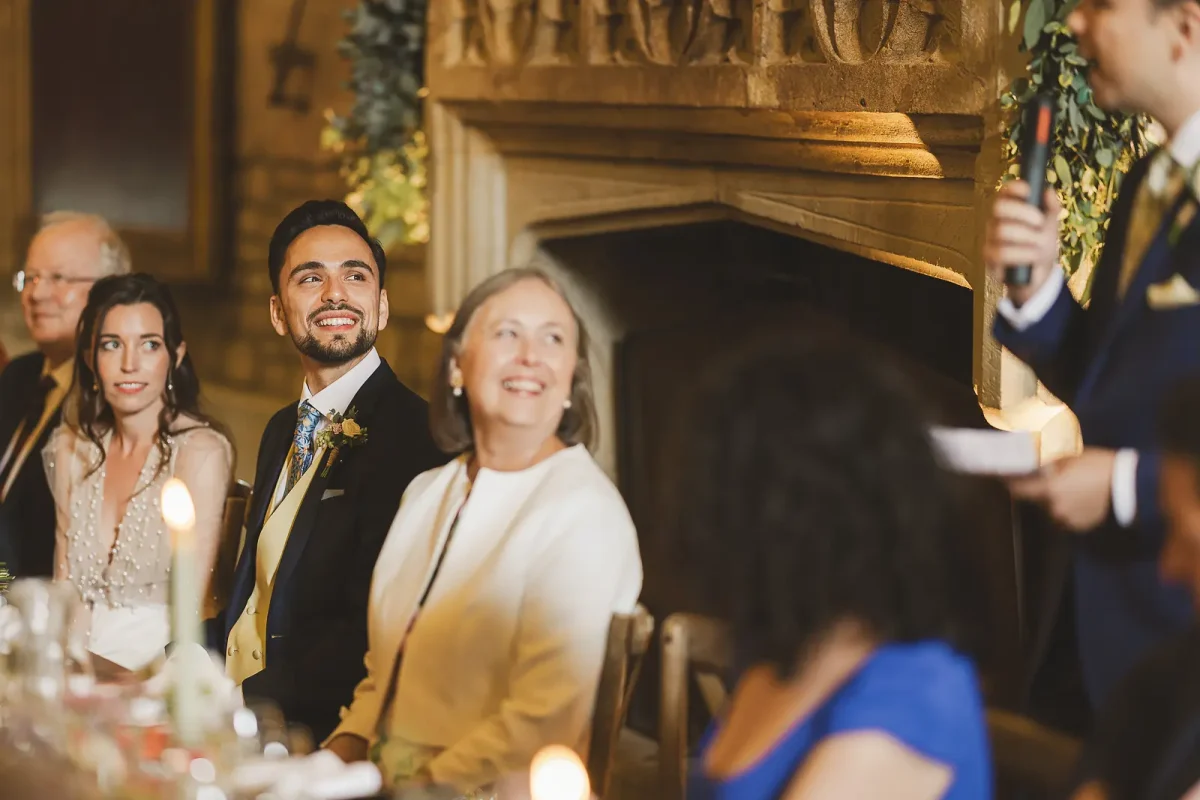 Bride and groom laughing and smiling at the top table during wedding speeches beside the stone fireplace in the tithe barn at Hinton St Mary Estate