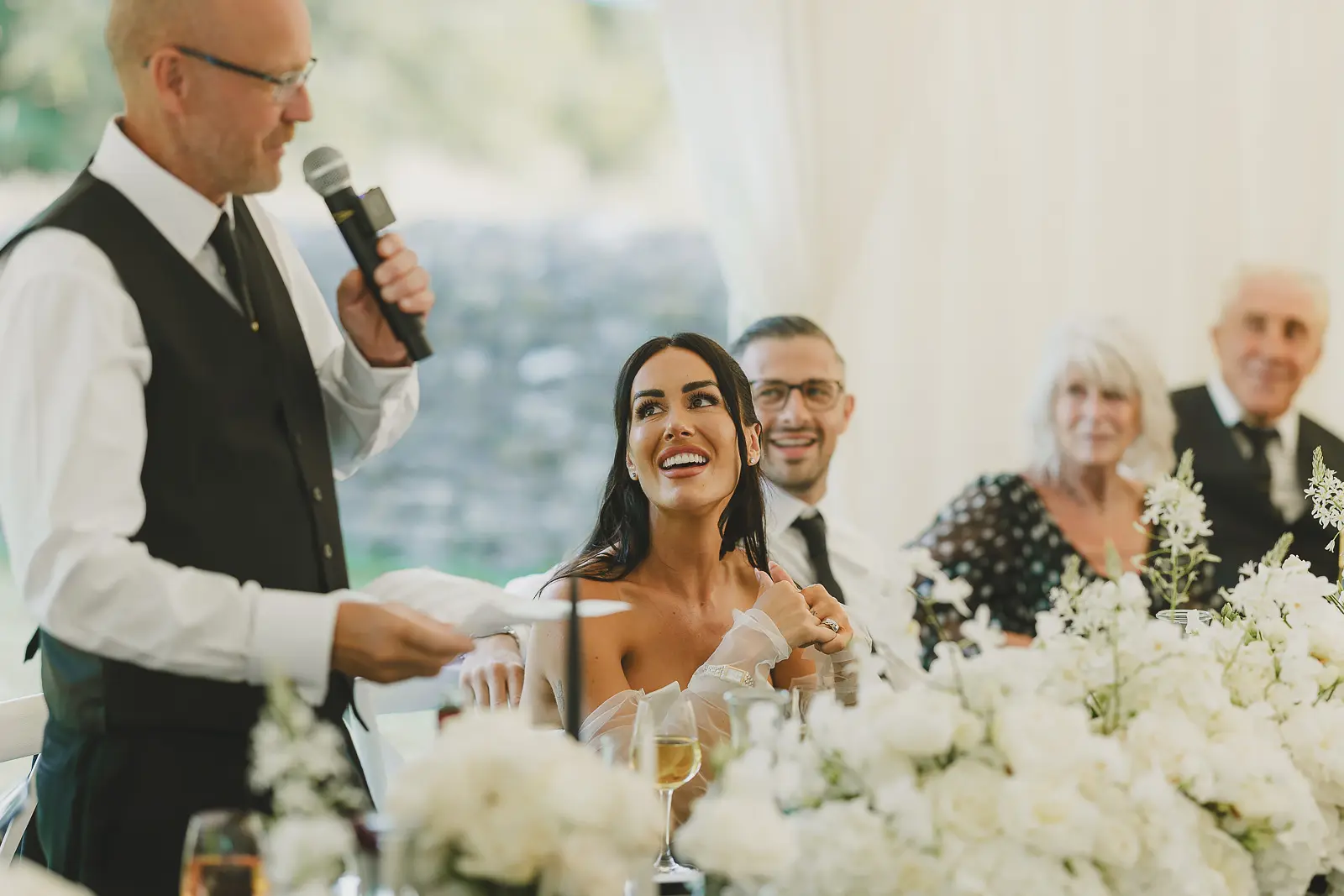 Bride laughing with delight at the top table during her father's wedding speech in the marquee, white florals in the foreground