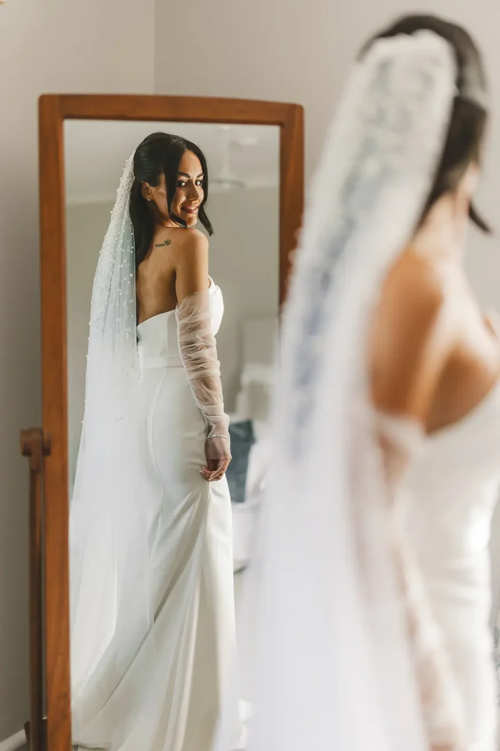 Bride looking at her reflection in a full-length mirror in her wedding dress with pearl-beaded veil, getting-ready morning portrait