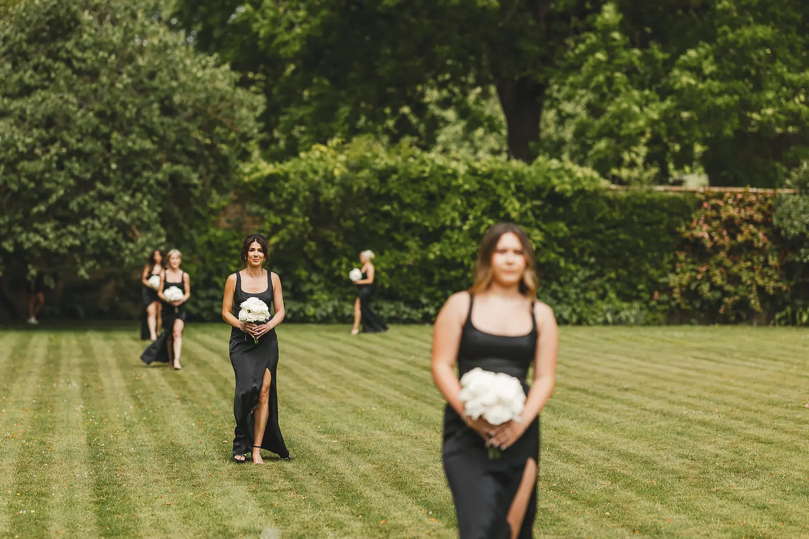 Four bridesmaids in black slit dresses carrying white bouquets processing across the striped lawn at Mapperton Gardens in summer
