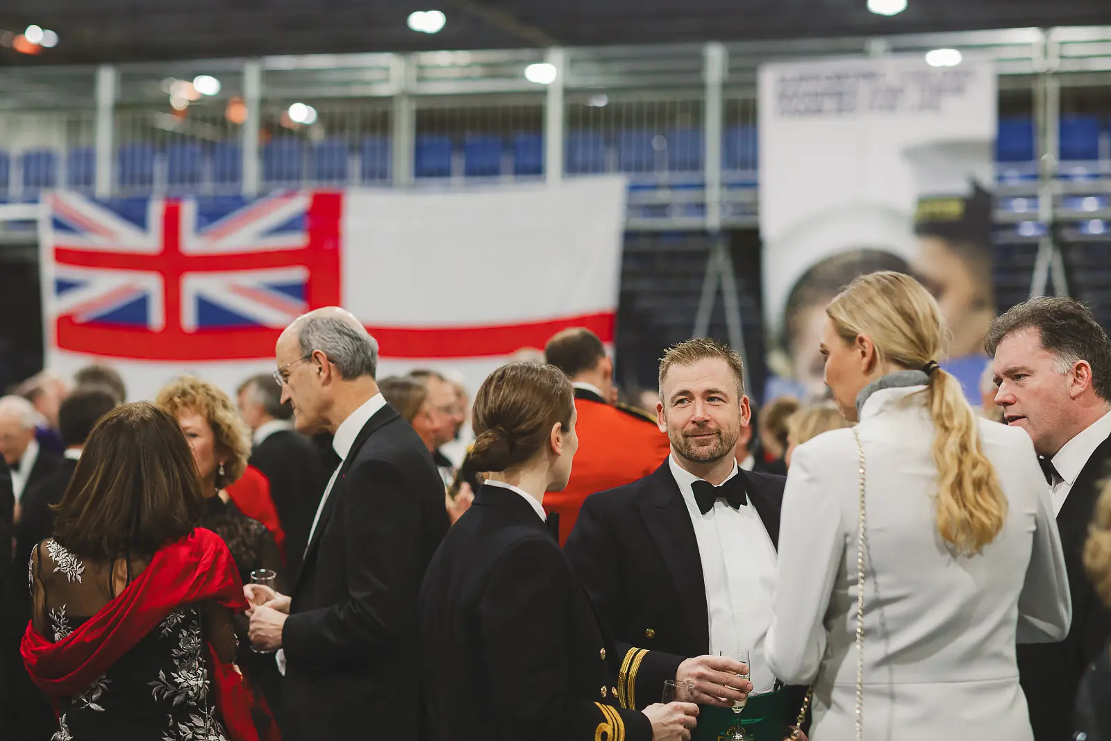 Busy drinks reception with mixed military and civilian guests in formal attire, blue tiered seating and White Ensign in background
