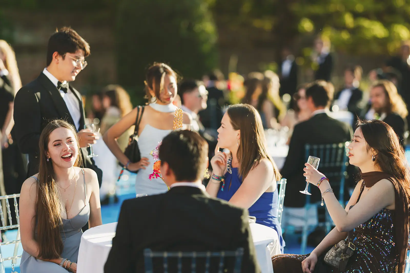 Candid moment of guests laughing and drinking champagne at outdoor tables during a summer ball reception