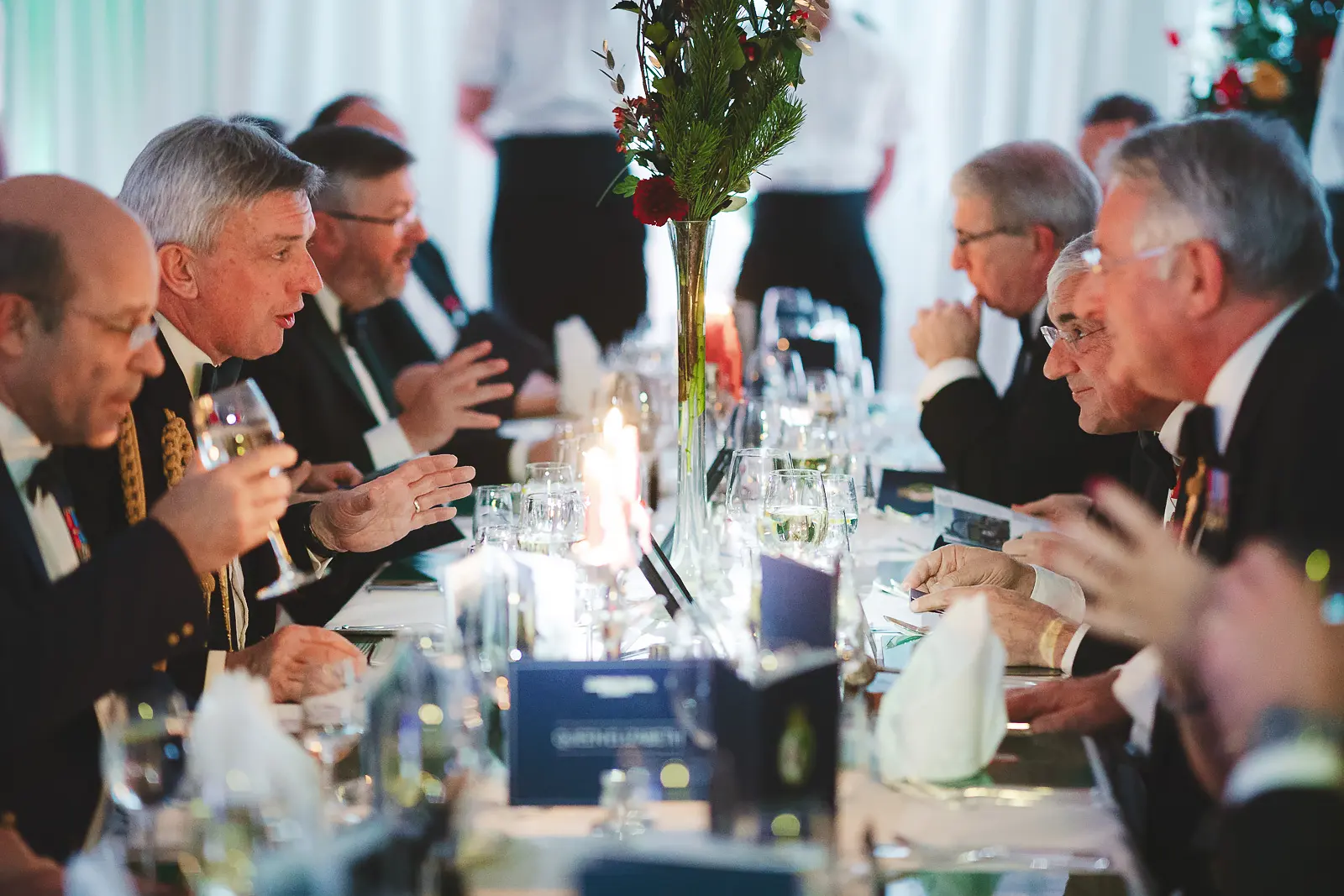 Guests in animated conversation at candlelit formal dinner table, tall floral centrepiece and wine glasses, documentary event photography