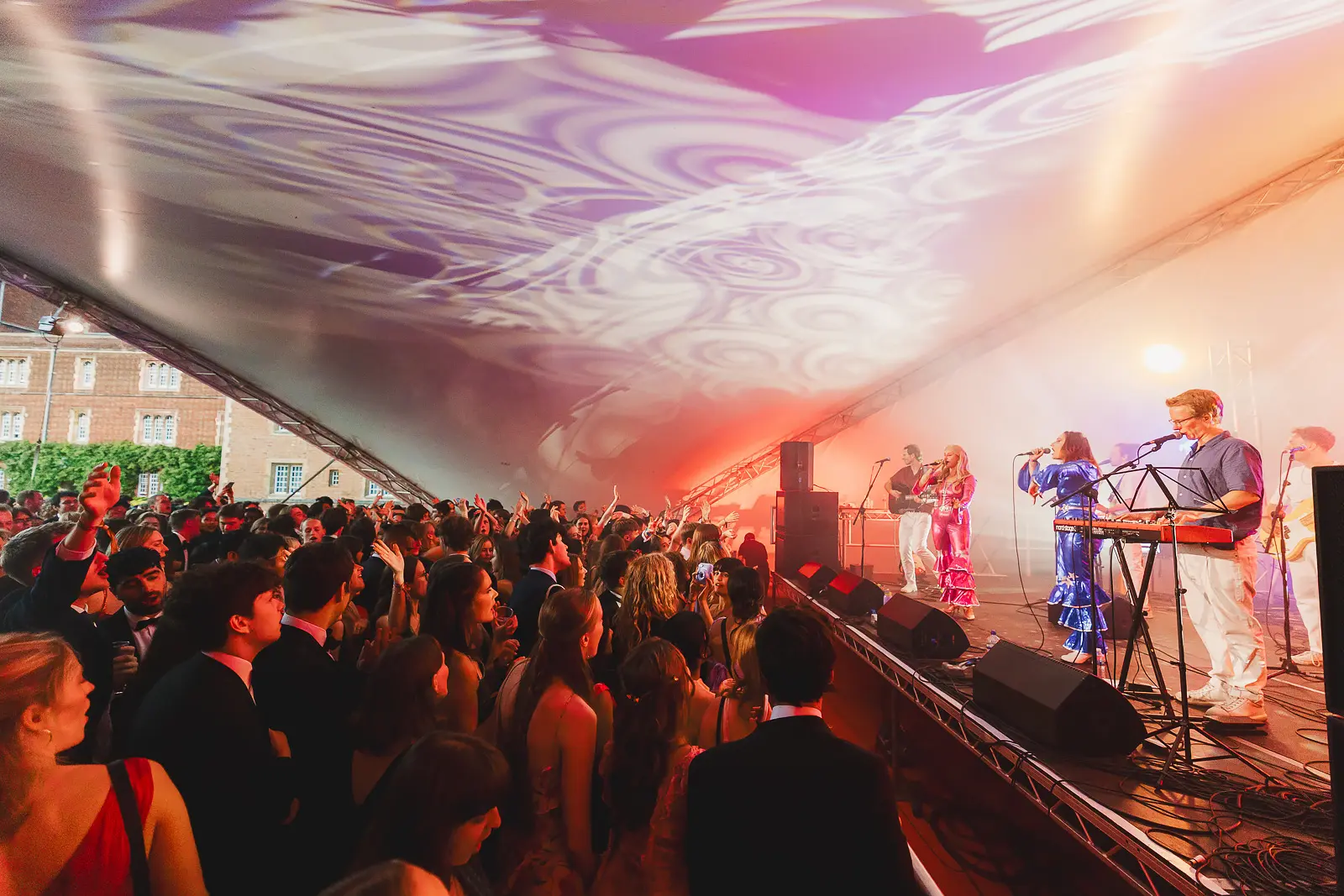 Live band performing on a canopy stage with psychedelic ceiling projection and a packed crowd at a summer ball