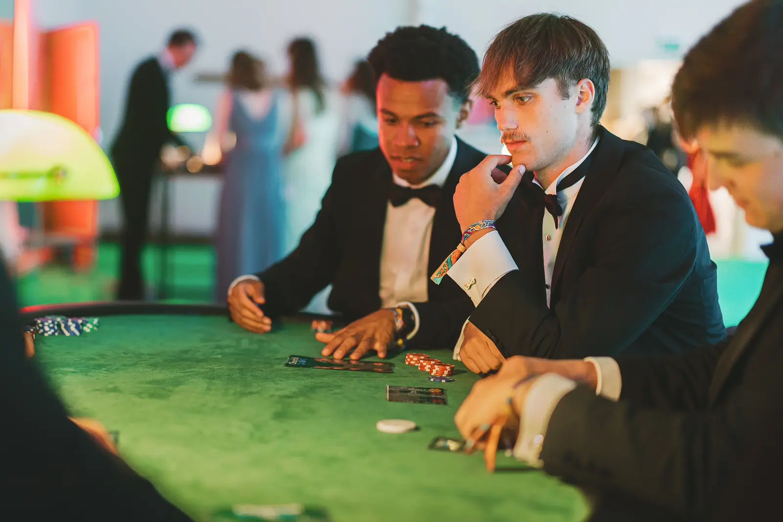 Guests in black tie at a casino table with poker chips under green lamp lighting at a summer ball