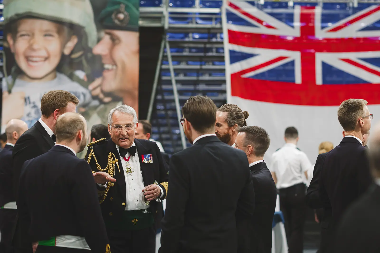 Senior officer in full ceremonial dress with sash and medals surrounded by black-tie guests on hangar deck, projected image in background