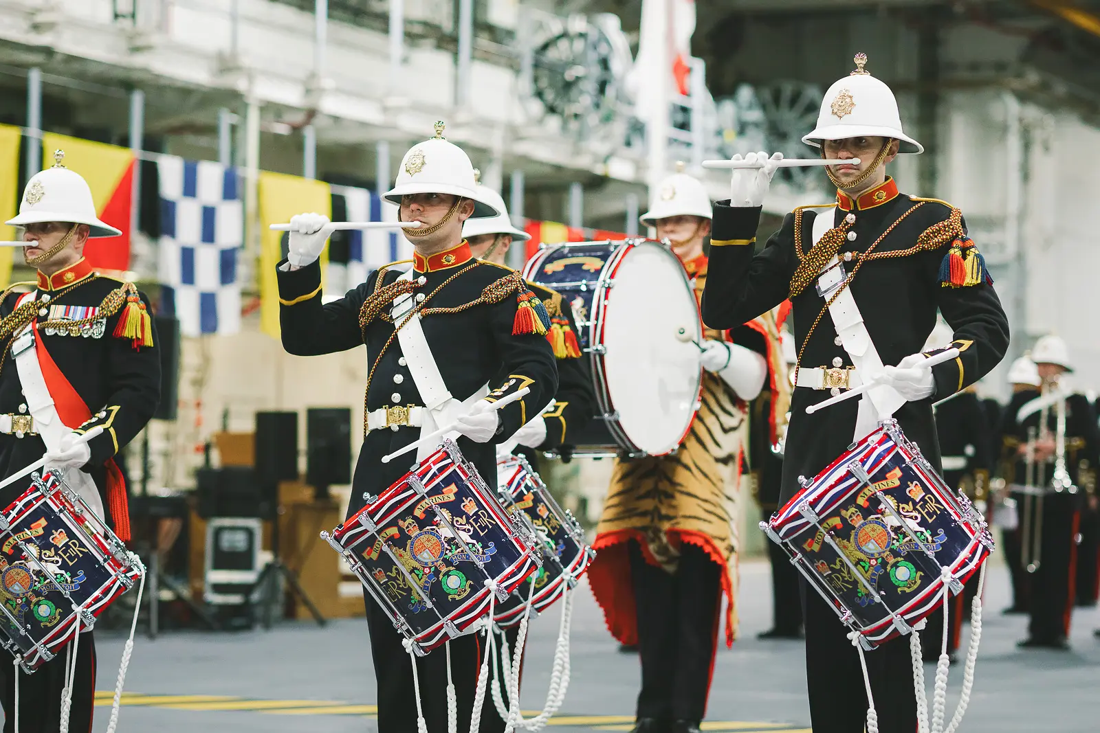 Ceremonial drummers in white pith helmets with drumsticks raised mid-beat, ornate decorated drum skins and tiger-skin bass drum visible