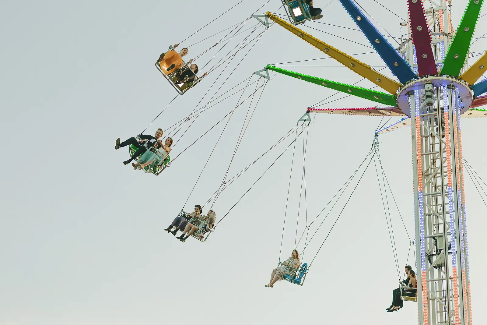 Formally dressed guests riding a chain-swing fairground attraction suspended against a pale evening sky at a Cambridge May Ball
