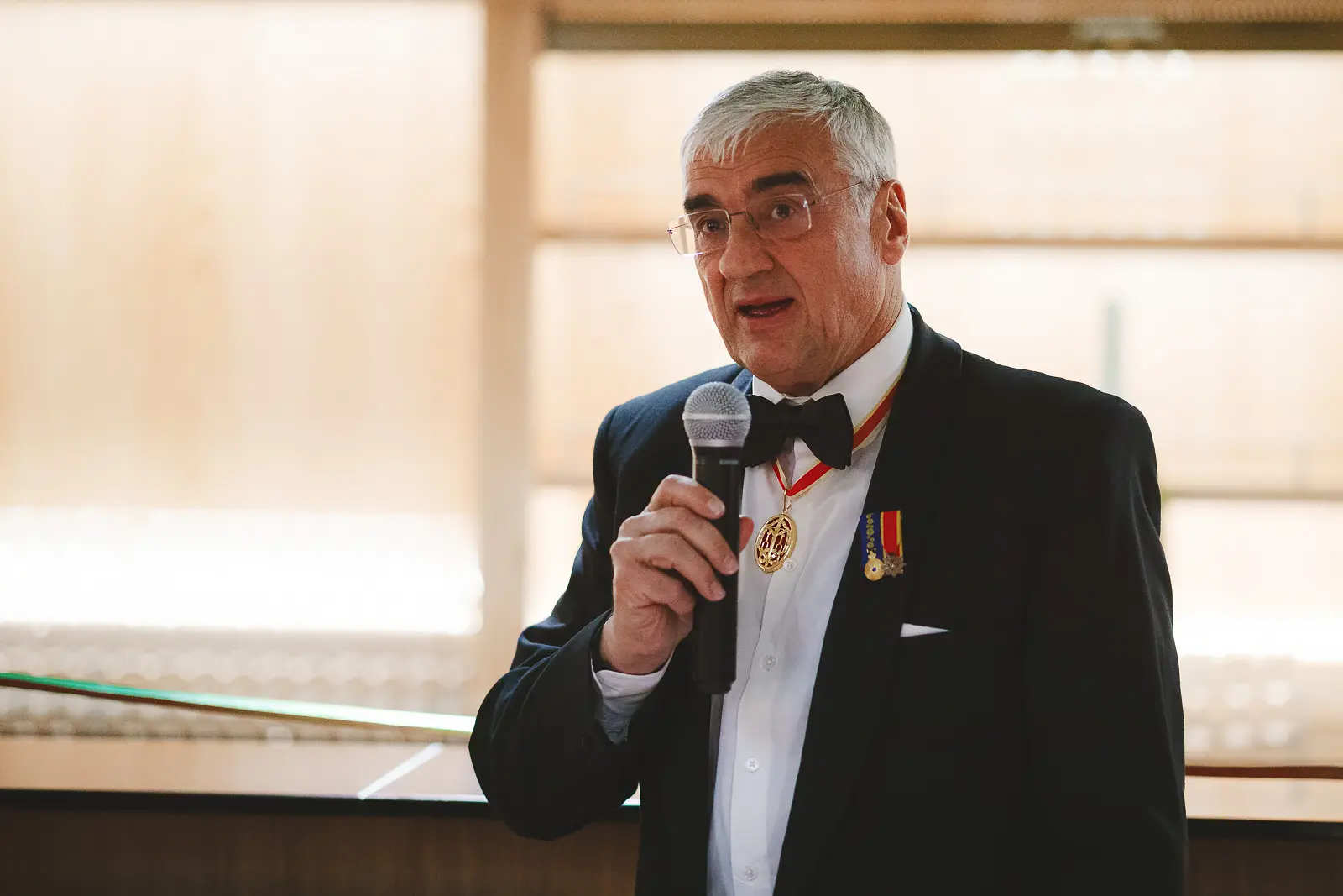 Civilian dignitary in black tie wearing gold medallion chain of office delivering speech into microphone at formal dinner event