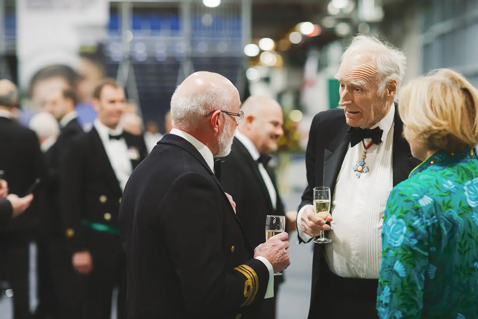 Naval officer and decorated dignitary in close conversation during formal reception holding champagne glasses, candid documentary photography