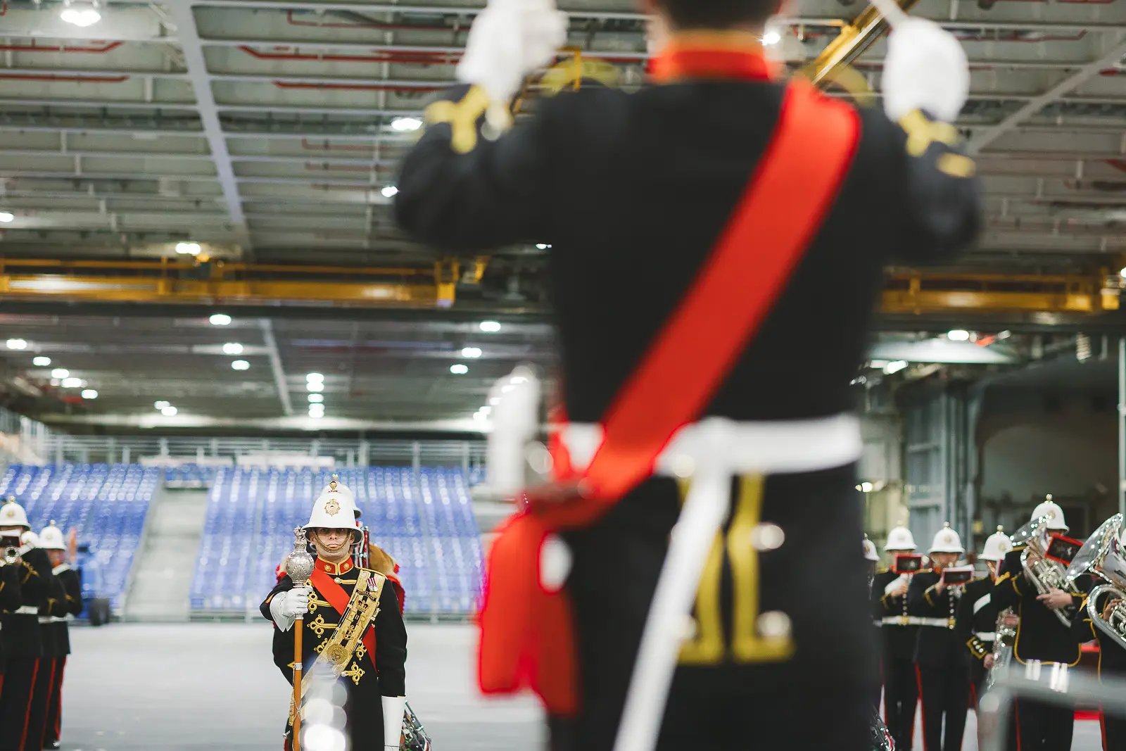 View past conductor with raised baton and red sash to drum major holding ceremonial mace during military band performance on hangar deck