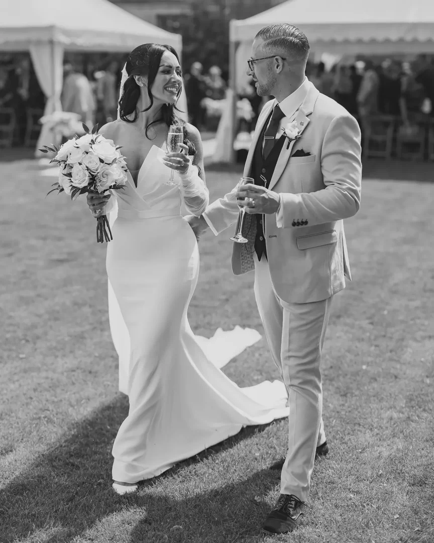 Black and white editorial portrait of the bride and groom clinking champagne flutes on the lawn after the ceremony at Mapperton