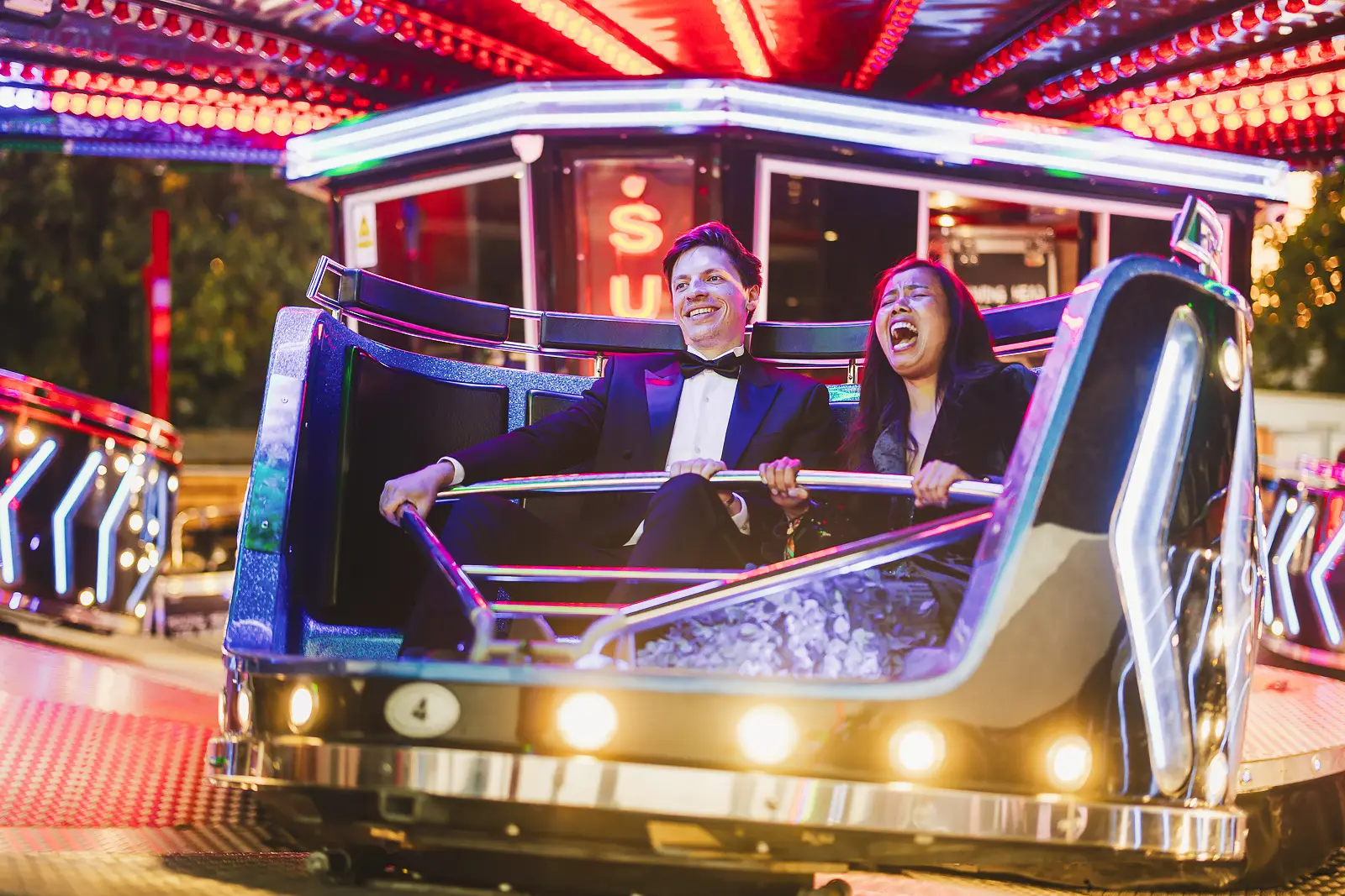 Couple in black tie laughing on a waltzers fairground ride surrounded by neon lights at a summer ball