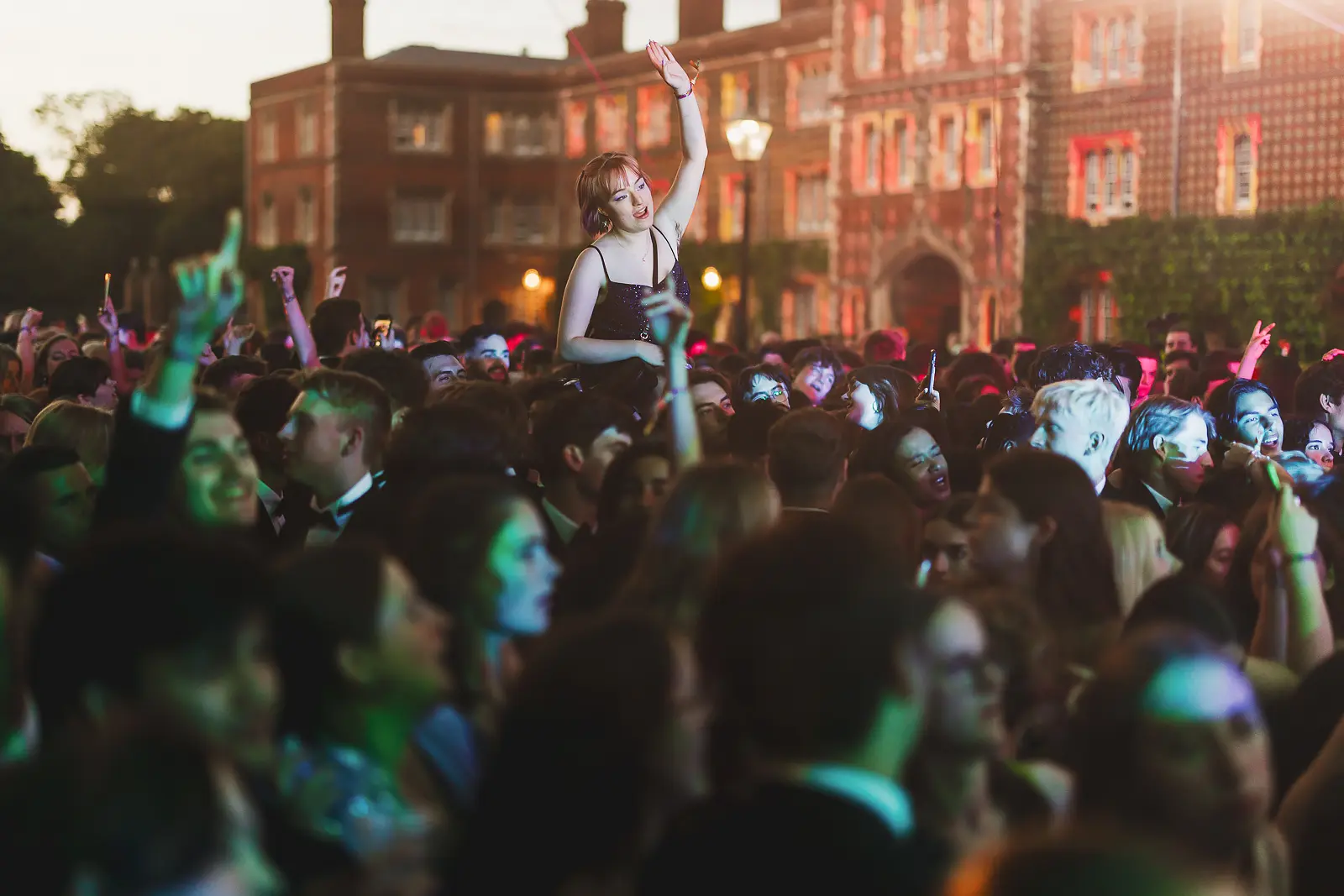 Guest in a sparkly dress lifted above the crowd with a lit college building behind at dusk during a summer ball