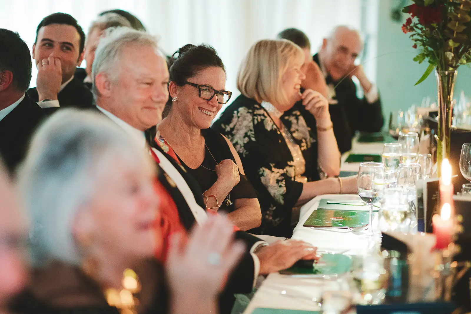Row of smiling formal dinner guests watching after-dinner entertainment, woman with glasses and decorated dignitary prominent, candid event photograph