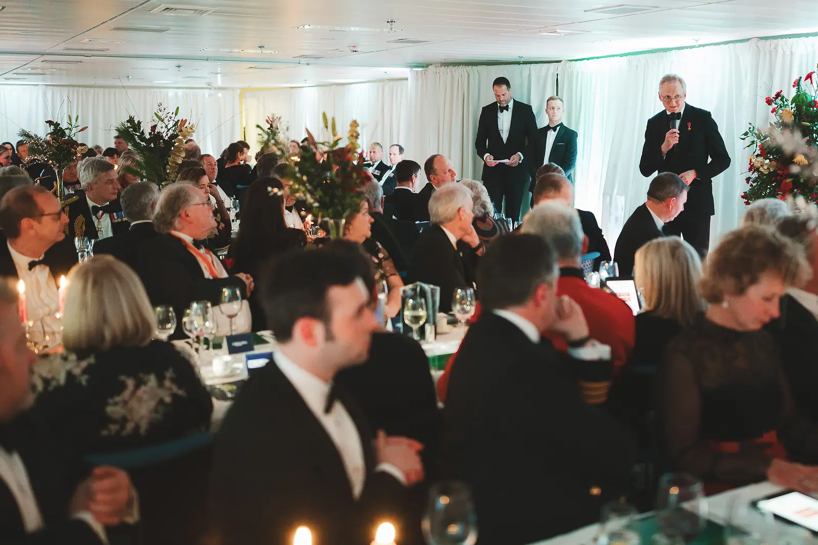 Wide view of full formal dinner room during speeches, speaker at far end with microphone, guests at candlelit tables with festive floral arrangements