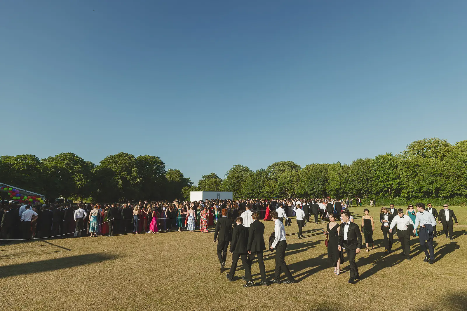 Wide documentary shot showing hundreds of guests in formal wear gathered on open grounds at a large-scale summer ball