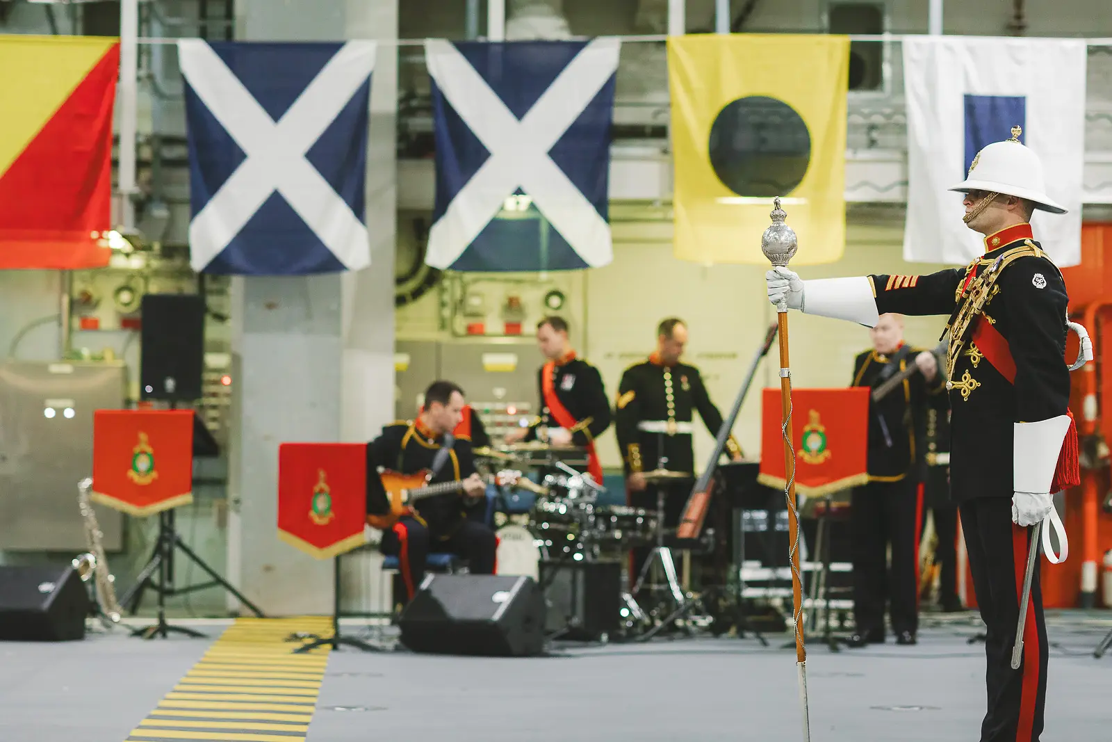 Drum major in ceremonial dress stands at attention with mace while evening band performs on stage behind, naval signal flags overhead