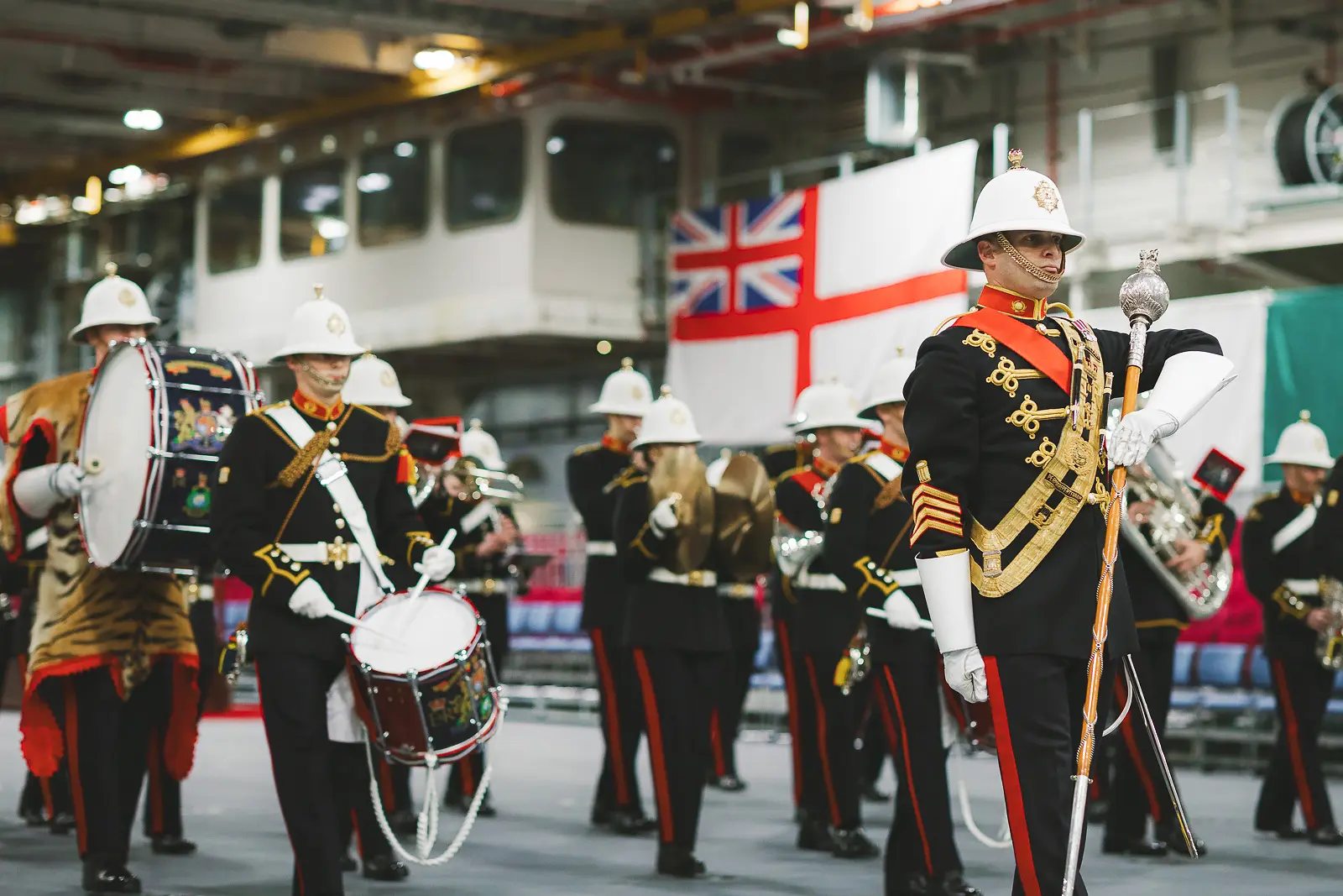 Drum major in ceremonial dress with gold braid and mace leads military band performance, White Ensign and Union flag in background