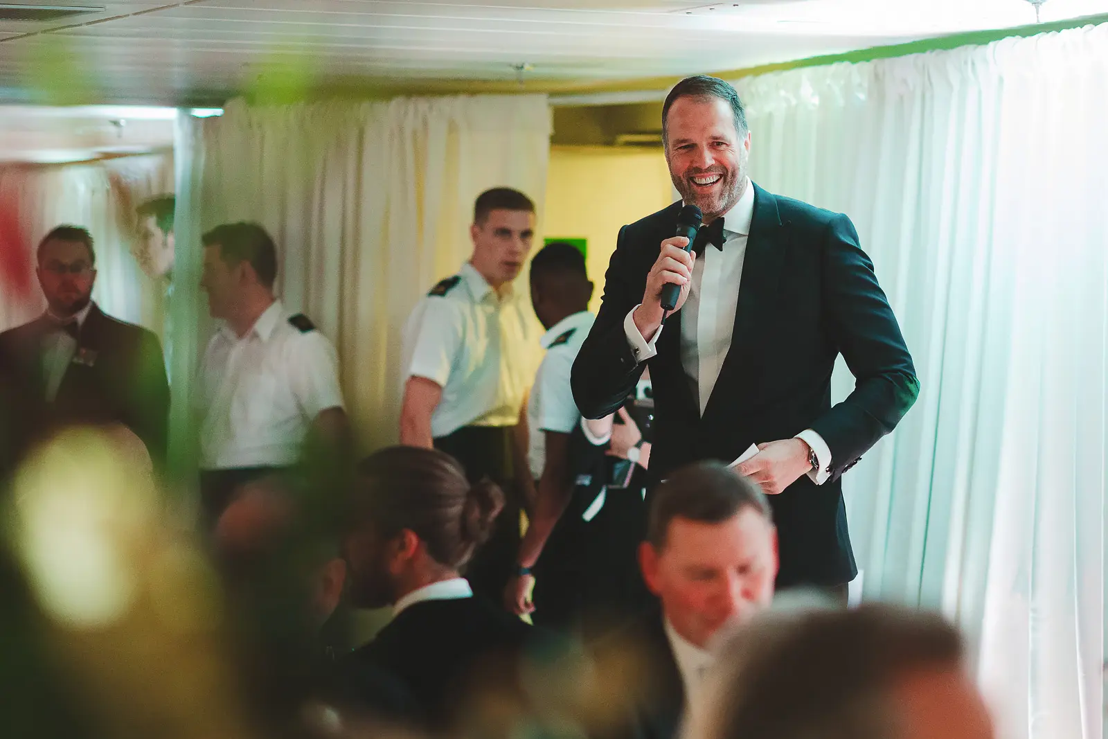 Laughing man in black tie holding microphone as after-dinner speaker or emcee entertaining guests, naval stewards visible in background