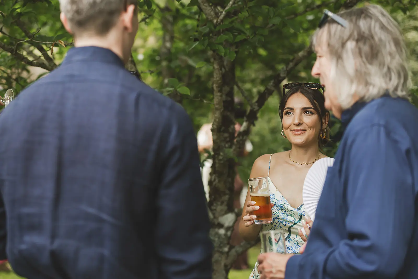 Young female guest in floral dress holding a drink and paper fan chatting with guests under trees at the outdoor drinks reception at Mapperton
