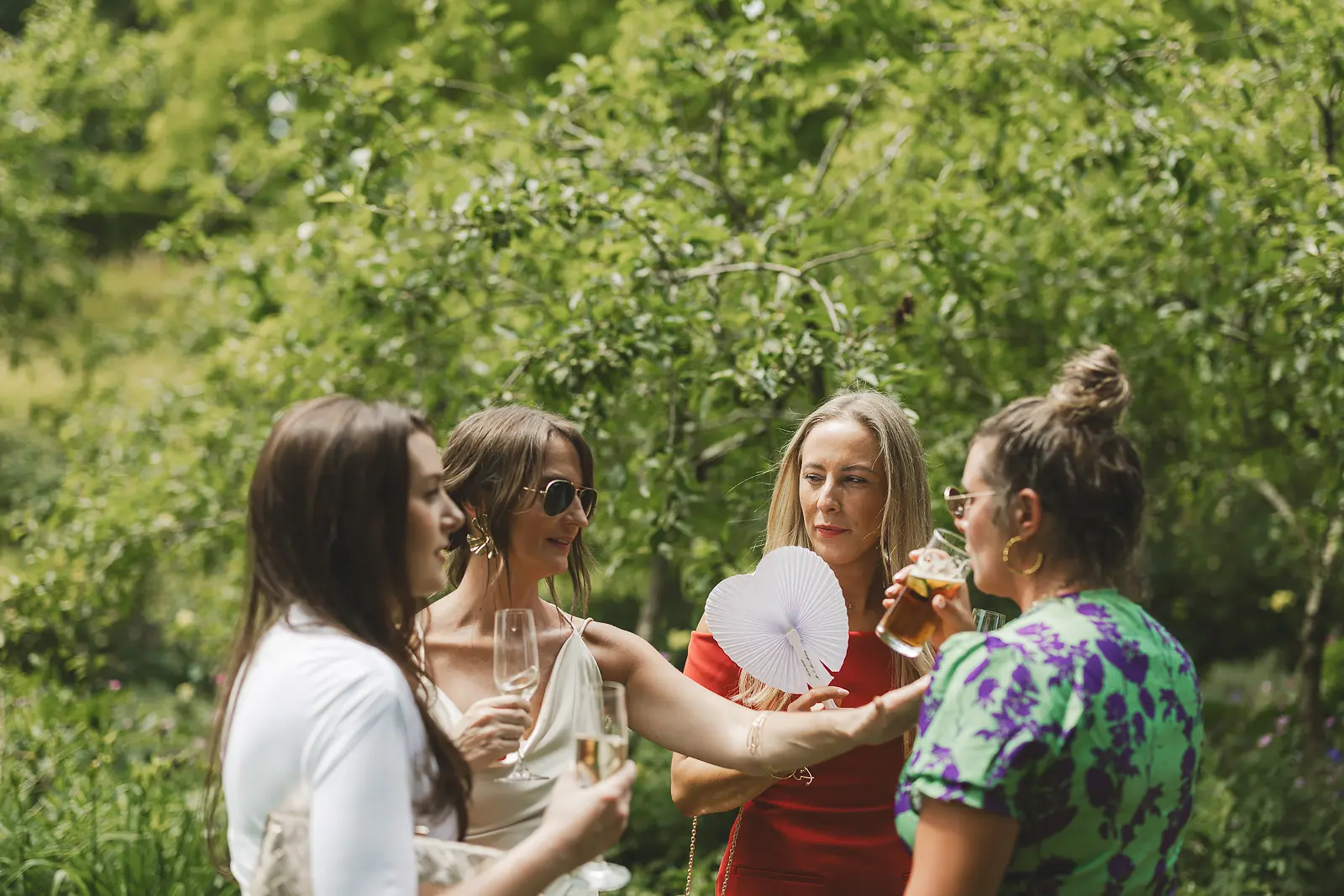 Female wedding guests enjoying the outdoor drinks reception in the garden at Mapperton, using paper fans in the summer heat